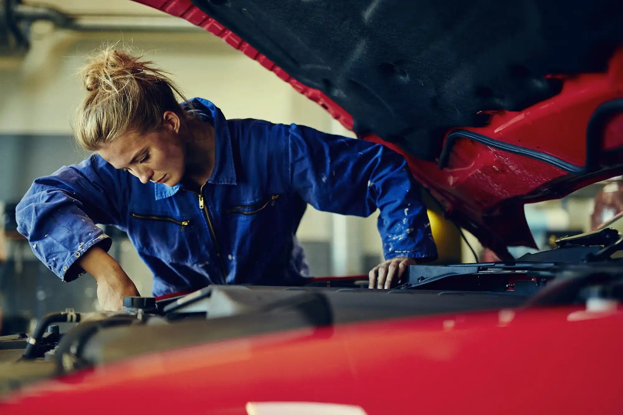 A mechanic in blue coveralls working under the hood of a red vehicle.